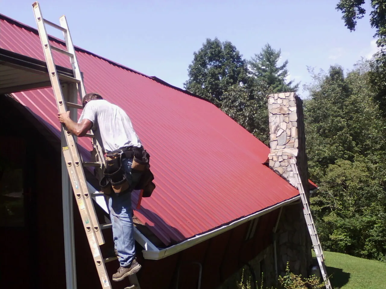 Roofer on a ladder beside a red metal roof and stone chimney — Soffit & Fascia Repair by Roofing Brothers in Faith